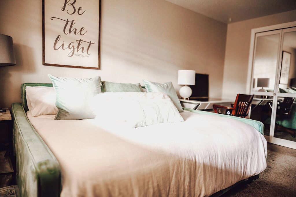Una cama bien hecha con cabecera verde, una mesita de noche con lámpara y una pared con una frase motivacional.