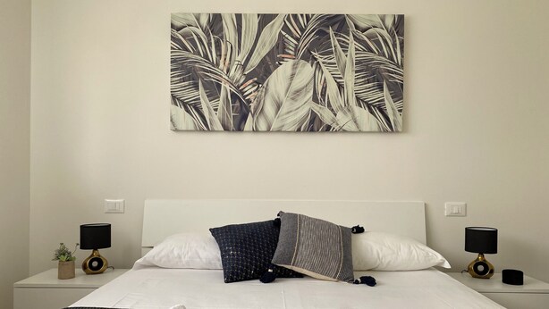 Een net opgemaakt bed met witte lakens, een donzig kussen met een patroon, en een kunstwerk van een tropisch blad aan de muur.
