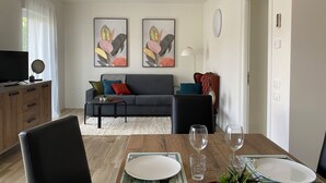 Een moderne woonkamer met een grijze bank, kleurrijke kussens en twee ingelijste bloemstillevens. Op de voorgrond staat een eettafel met borden en glazen.
