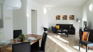 A modern living room with a black sofa, a coffee table, and a dining area with a wooden table and chairs. There are three paintings on the wall and a floor lamp.