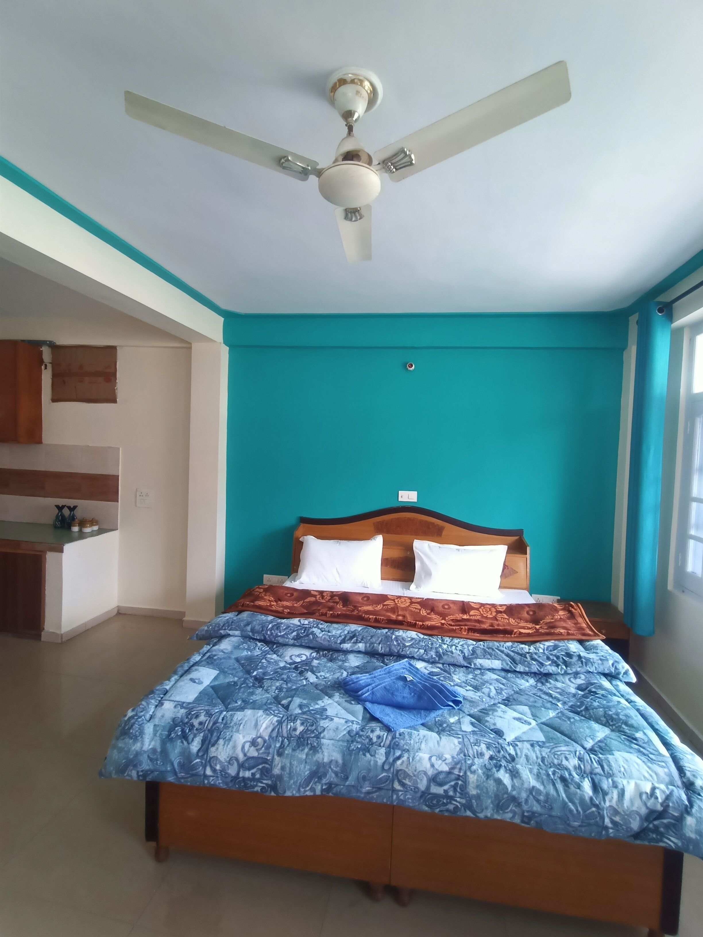 A bedroom with a bed, a ceiling fan, a window, and a kitchenette area.
