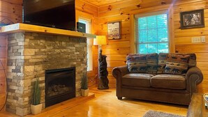 A cozy cabin interior with a stone fireplace, a leather sofa, and a mounted television. - Gatlinburg, Charming Cabin Hearth of Nature With Pool Table, Fireplace, Hot Tub (Sevierville)