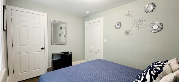 Quarto com duas portas brancas, uma cama com colcha azul, cômoda preta e decorações nas paredes.