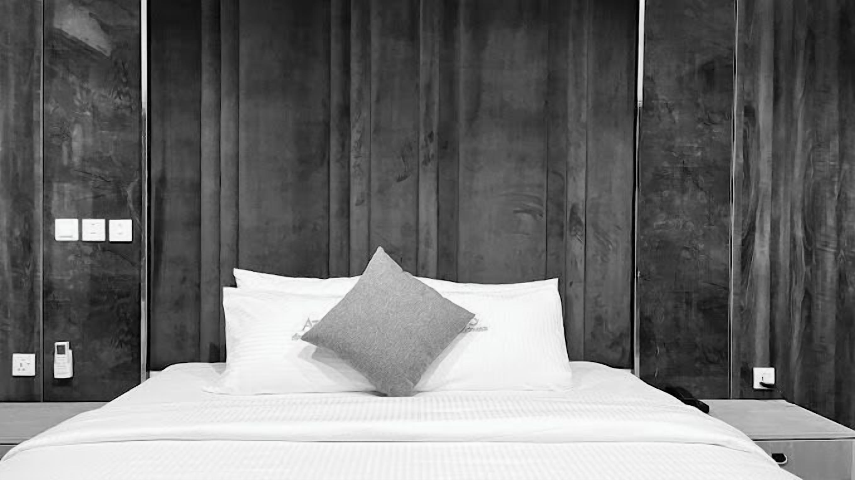 Una cama con ropa de cama blanca y una almohada gris, frente a un panel de pared de madera.