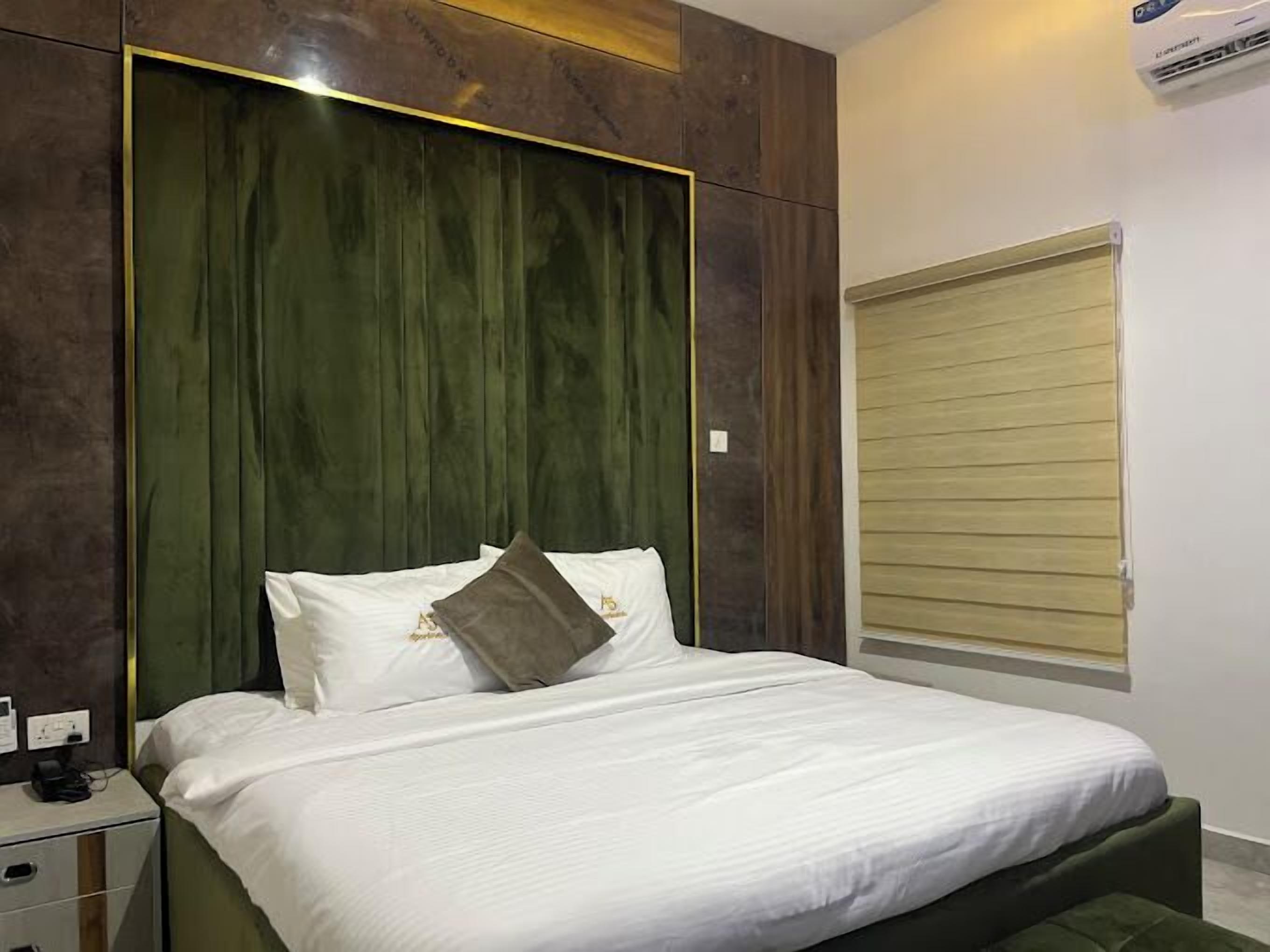 A neatly made bed with white linens and two pillows, a bedside table with a phone and charger, and a window with closed blinds.