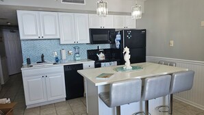A modern kitchen with white cabinets, a black refrigerator, a microwave, and a blender. There are two bar stools and a decorative piece on the island. - SEPT DEAL 1BR OCEANFRONT KING,pools,hotub,pristine,free parking & stunning views (Myrtle Beach)