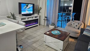 A modern living room with a flat-screen TV, a fireplace, a dining area, and a balcony with outdoor furniture. - SEPT DEAL 1BR OCEANFRONT KING,pools,hotub,pristine,free parking & stunning views (Myrtle Beach)