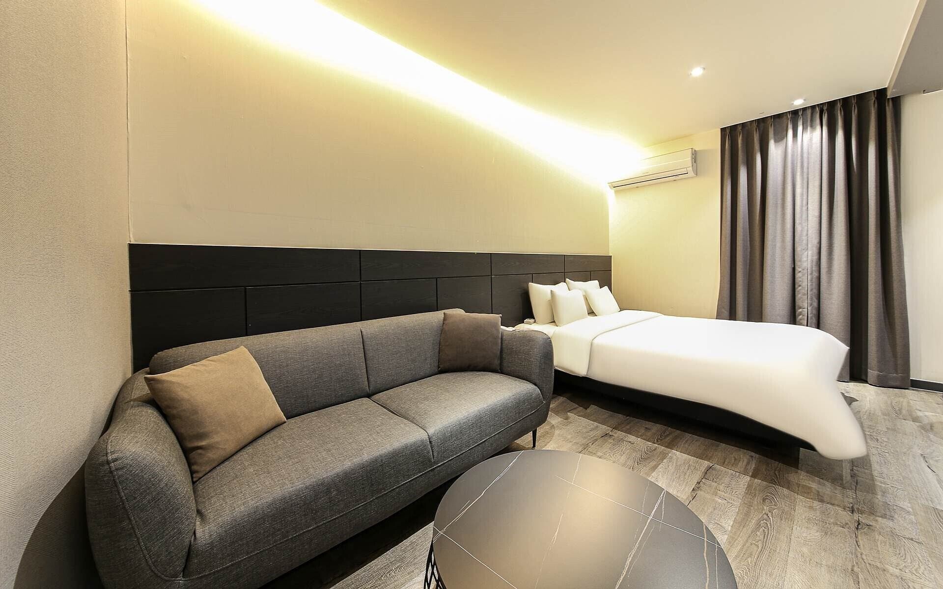 A modern hotel room with a grey sofa, a round black coffee table, a bed with white linens, and a dark headboard.