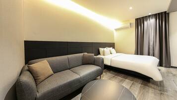 A modern hotel room with a grey sofa, a round black coffee table, a bed with white linens, and a dark headboard.