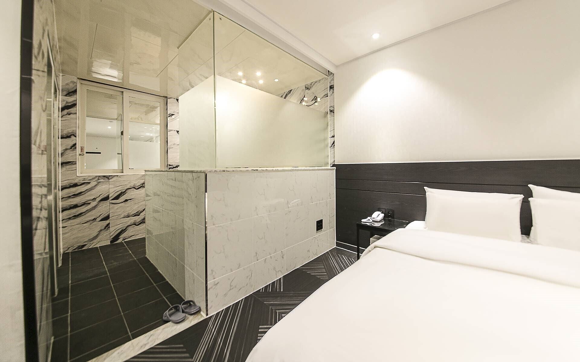 Una habitación de hotel moderna con una cama grande, una mesita de noche con teléfono y un área de ducha con paredes de vidrio.