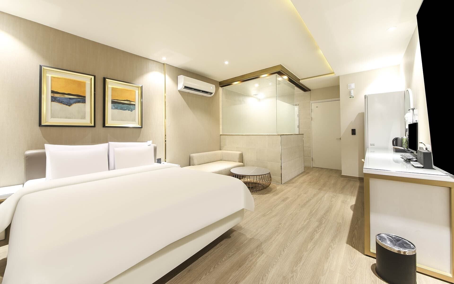 A hotel room with a large bed, a small sofa, a round table, and a desk with a computer and phone. There are two framed pictures on the wall, an air conditioning unit, and a trash can.