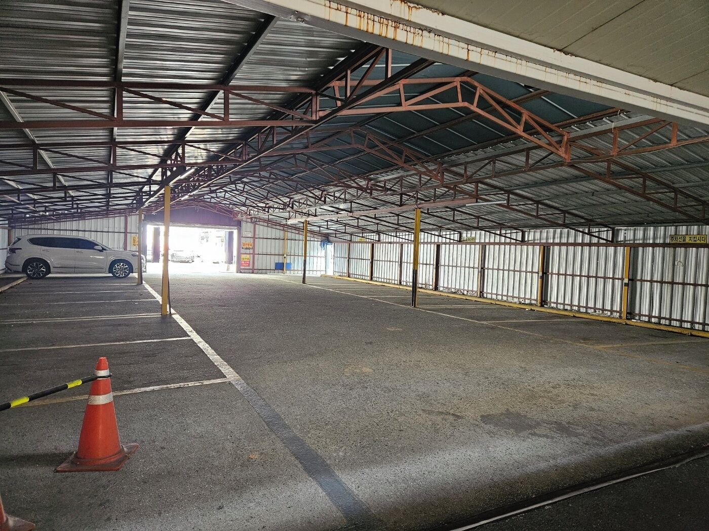 An indoor parking area with a white car parked in the foreground, a traffic cone, and a metal fence along the walls.