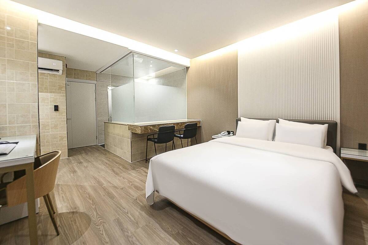 Habitación de hotel moderna con una cama grande, un escritorio con dos sillas, cabina de ducha de vidrio y una mesita de noche con teléfono.