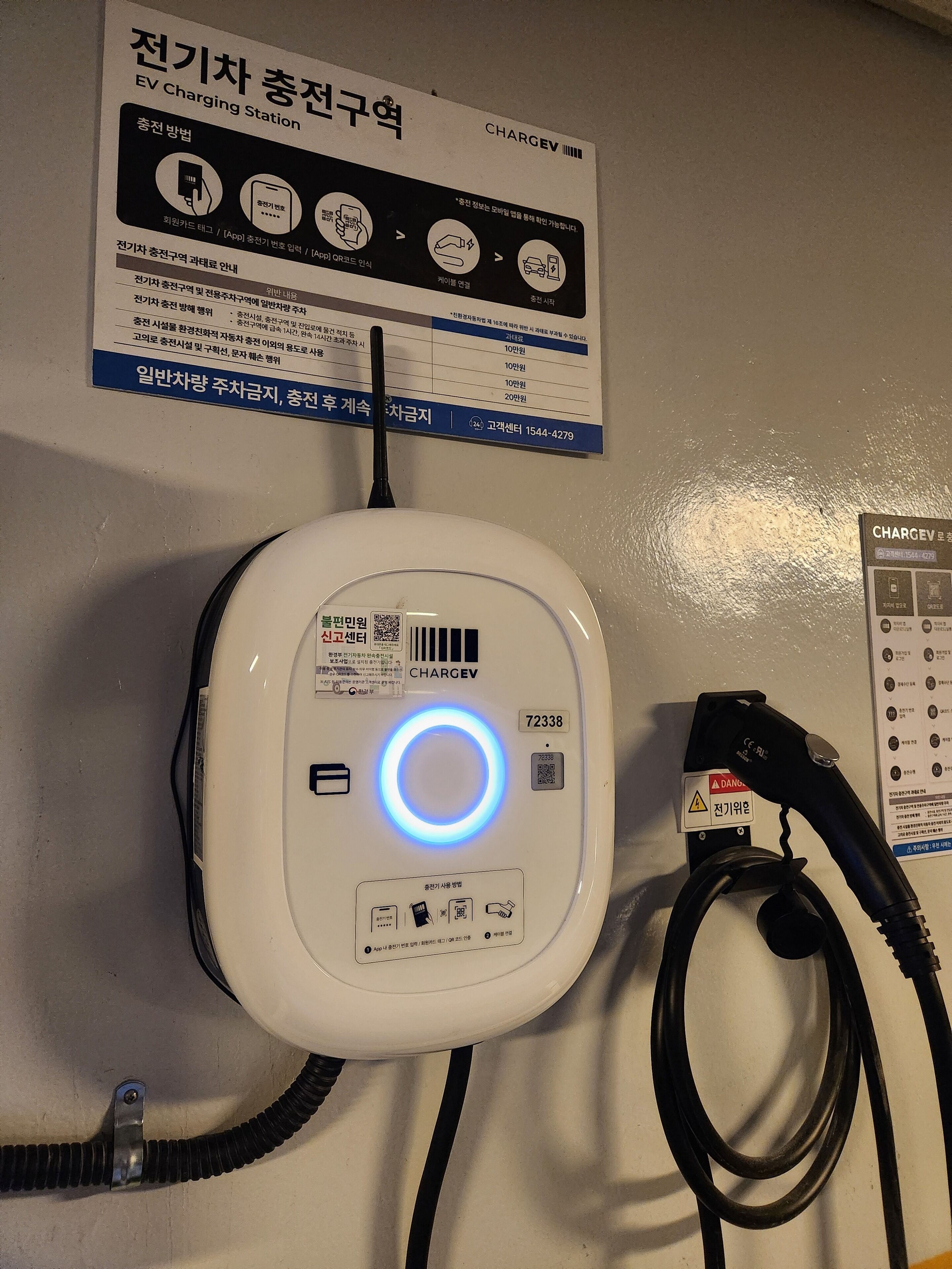 Una estación de carga para vehículos eléctricos blancos con un indicador luminoso azul, un cable de carga y un letrero encima.