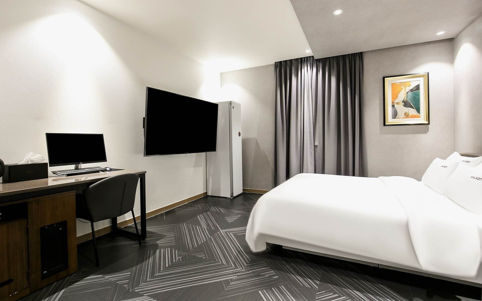 A hotel room with a bed, a desk with a computer, a flat-screen TV, and a refrigerator.