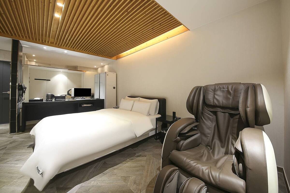 Habitación de hotel moderna con cama, sillón de masaje, escritorio con computadora y espejo.