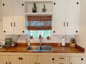 Une cuisine avec des meubles blancs, un plan de travail en bois, un évier en acier inoxydable et une fenêtre avec des stores.