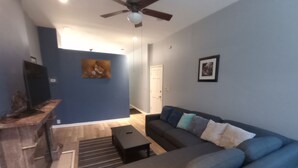 A living room with a dark blue wall, a gray sofa with multiple cushions, a wooden coffee table, a black TV stand, a ceiling fan, and a framed picture on the wall. - Beautiful 2 Bedroom located in a Unique Private Park. Near Beaches Near Downtown (Chicago)