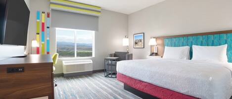 Una habitación de hotel con cama, escritorio, silla, televisor, ventana con vistas y un diseño estampado en la pared.