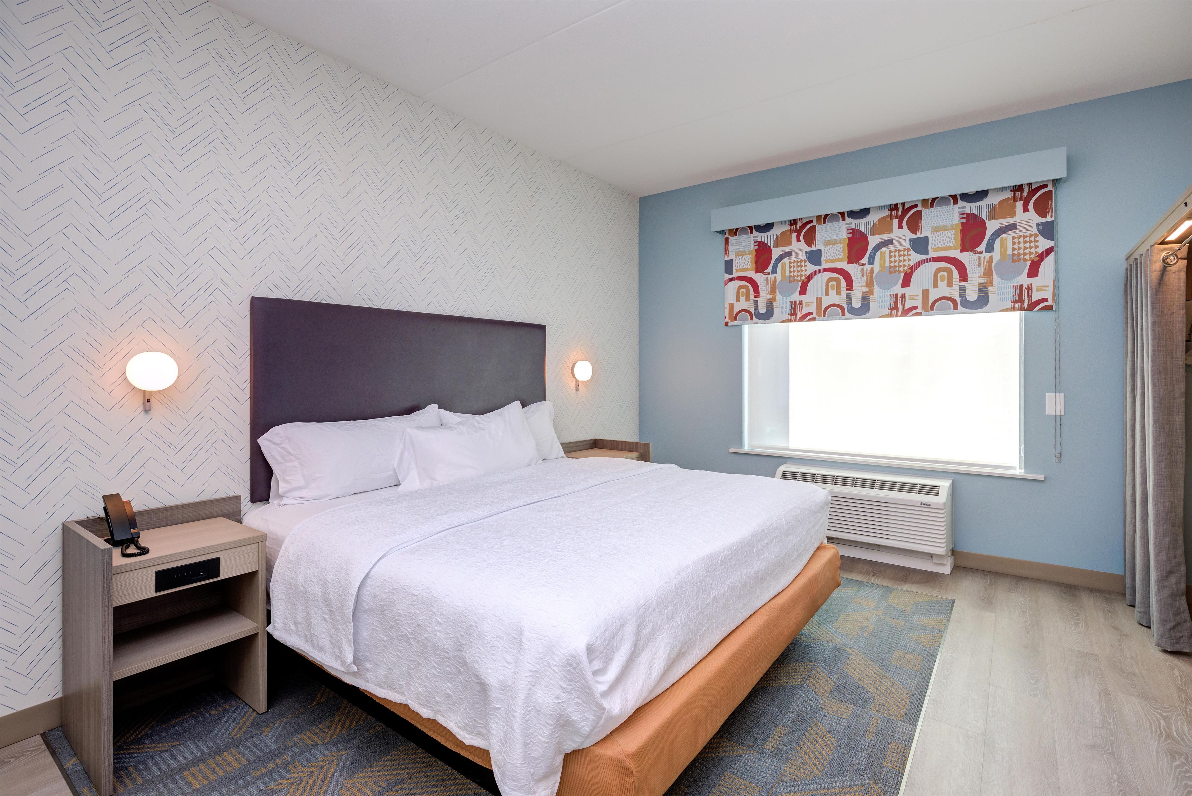 A bedroom with a bed, bedside table, lamp, and a window with a curtain.