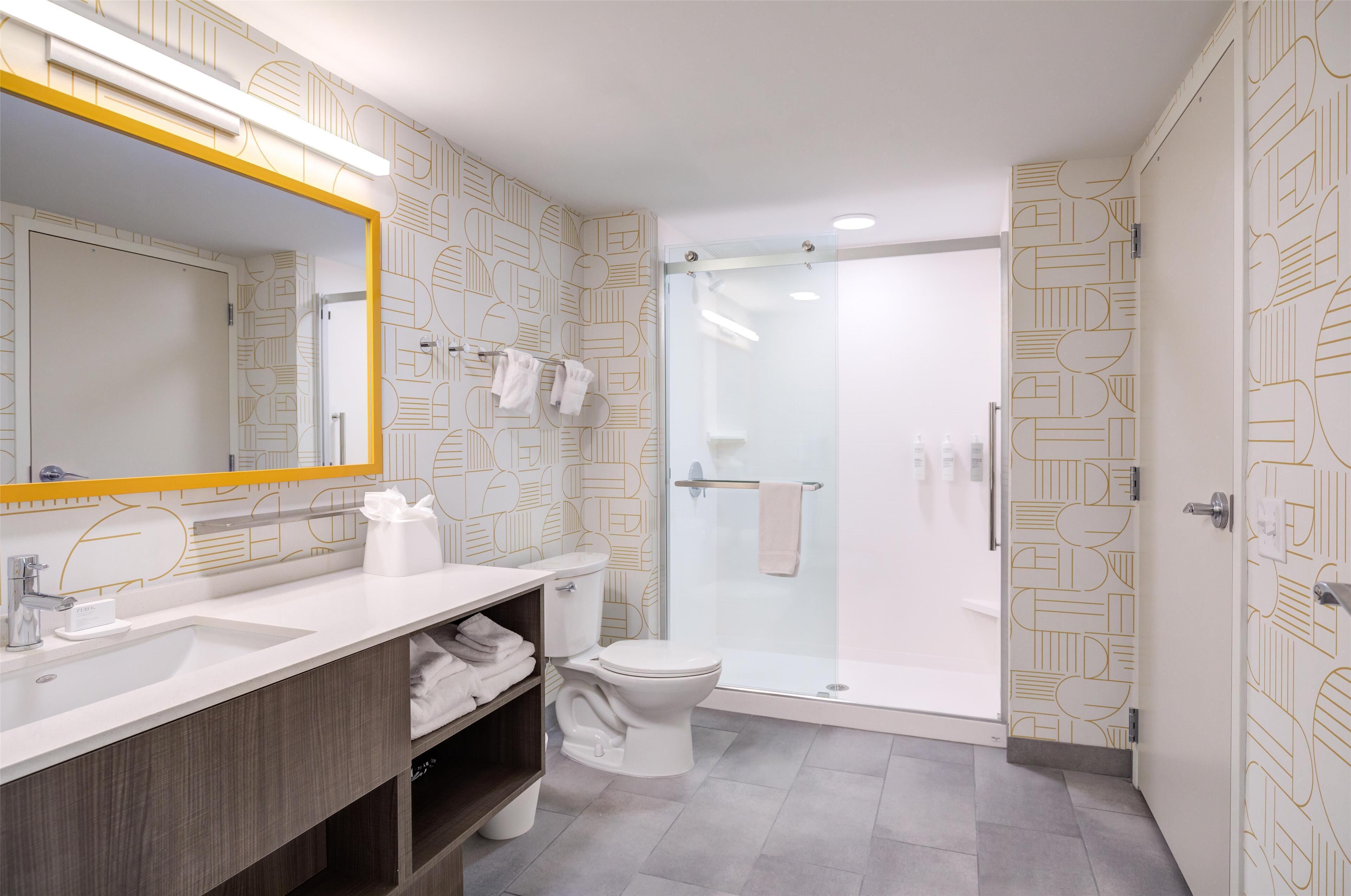 Une salle de bain moderne avec un grand miroir, des éléments d’ameublement blancs et un papier peint à motifs.