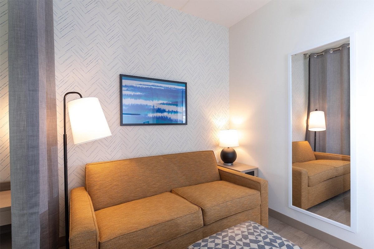 Una camera d'albergo moderna con divano, due abat-jour, uno specchio grande e una parete con motivo a spina di pesce.