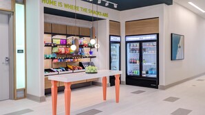 A snack display area with a sign reading "HOME IS WHERE THE SNACKS ARE" and a table with a fruit bowl. - Home2 Suites By Hilton Smithfield Selma (Smithfield)