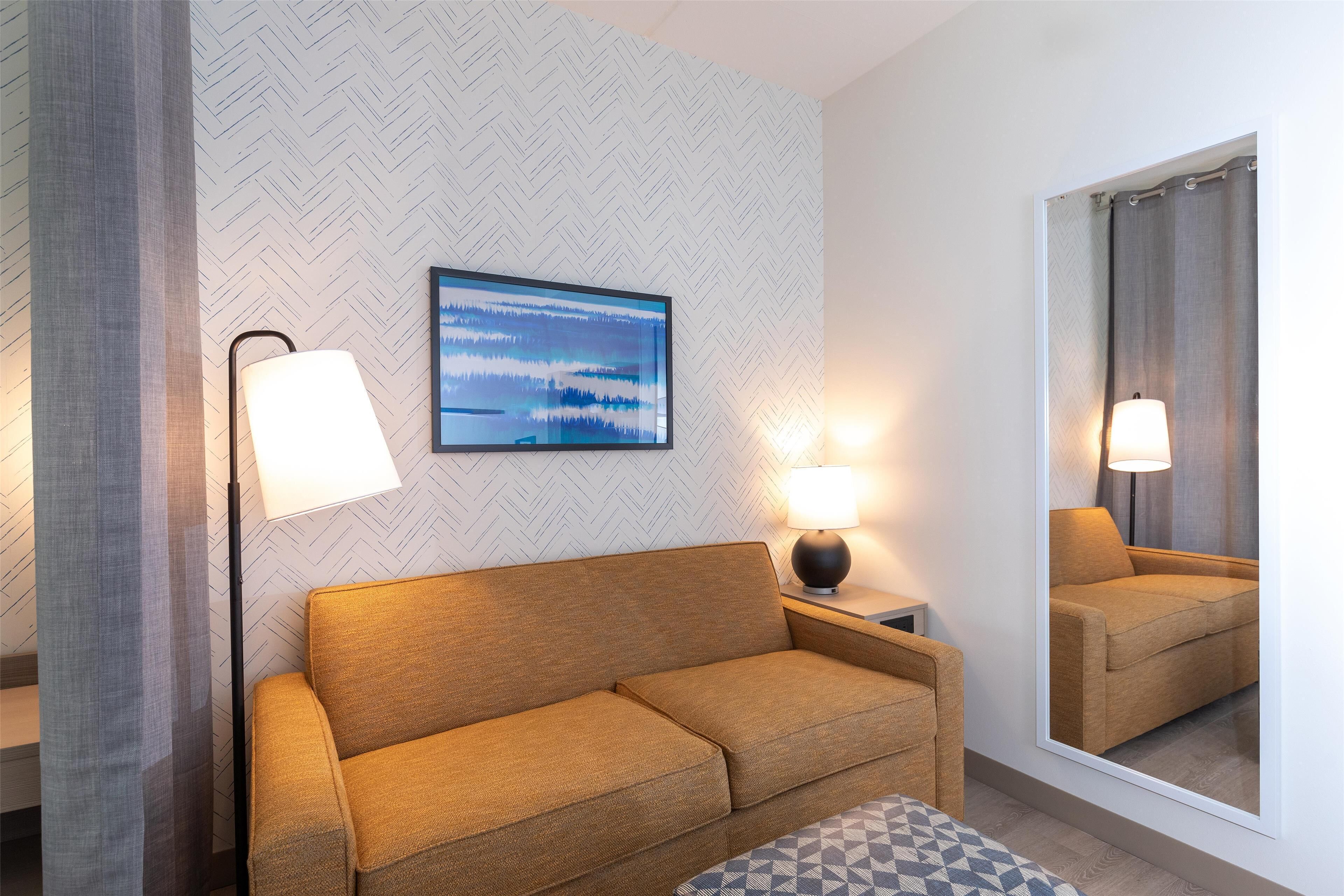 A modern hotel room with a sofa, two bedside lamps, a large mirror, and a herringbone-patterned wall.