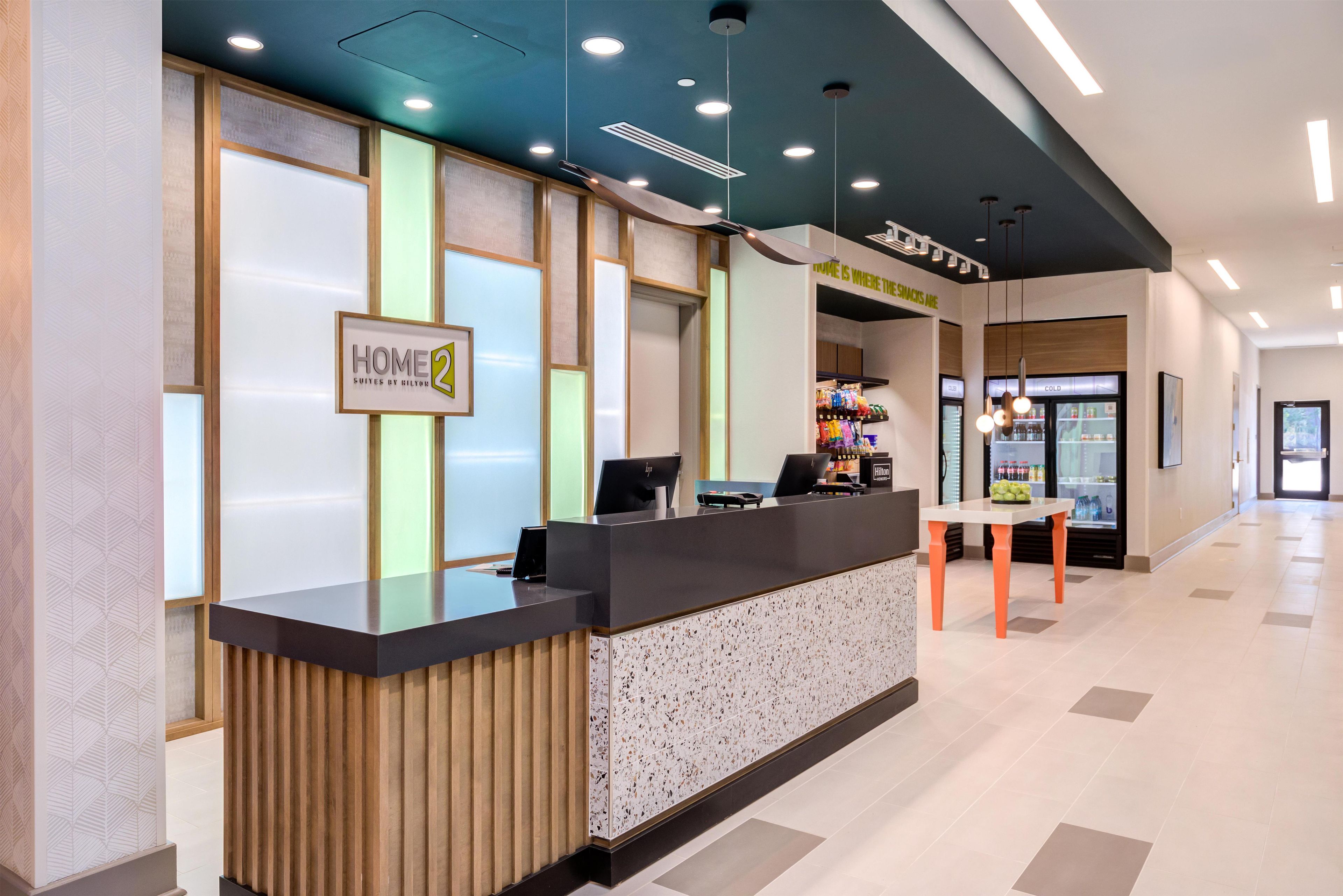 A hotel reception area with a modern design, including a reception desk, a sign reading 'HOME 2', and a 'COLD' beverage refrigerator.