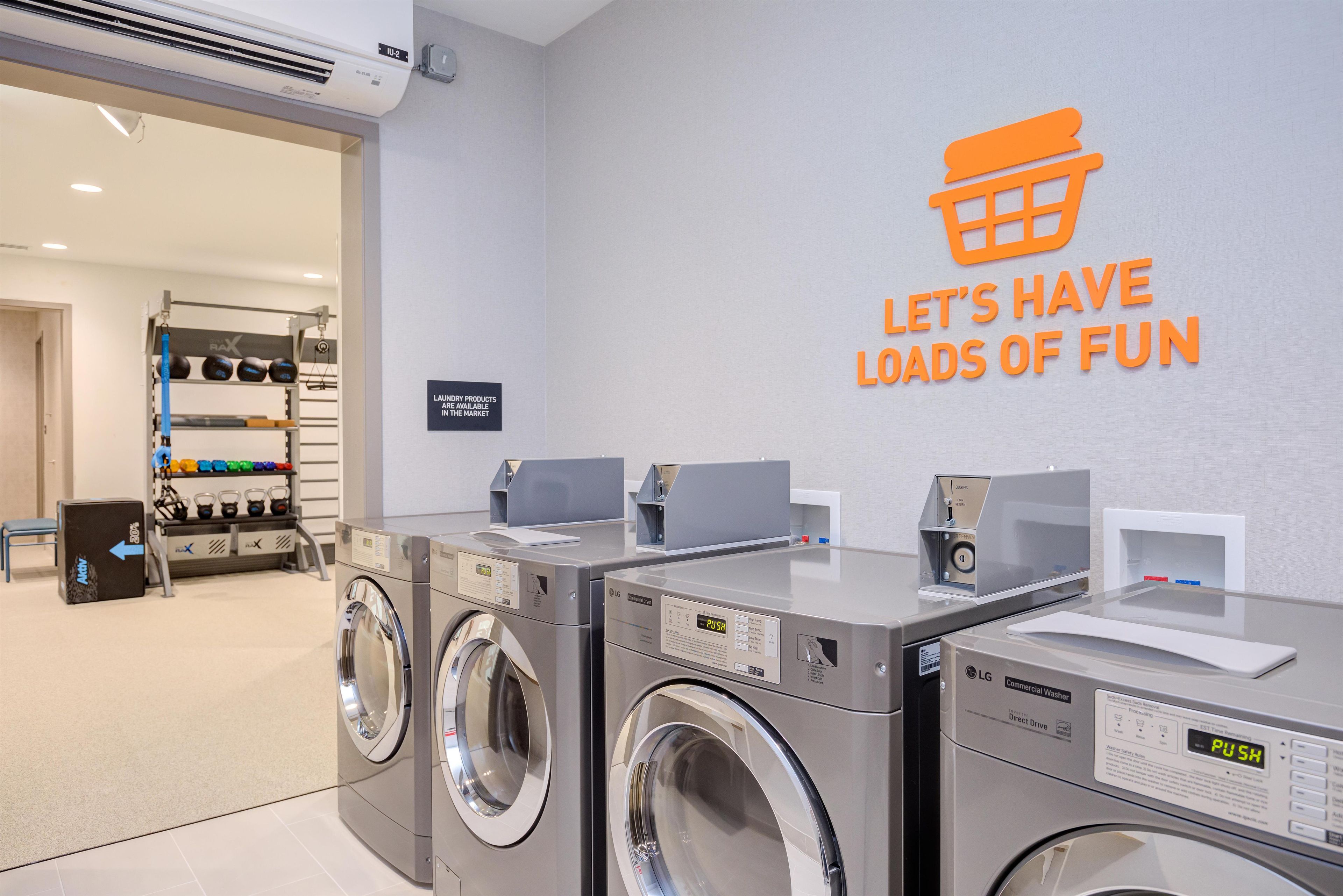 A laundromat with commercial washers and dryers, a sign indicating laundry products are available, and a wall sign with a message about having loads of fun.