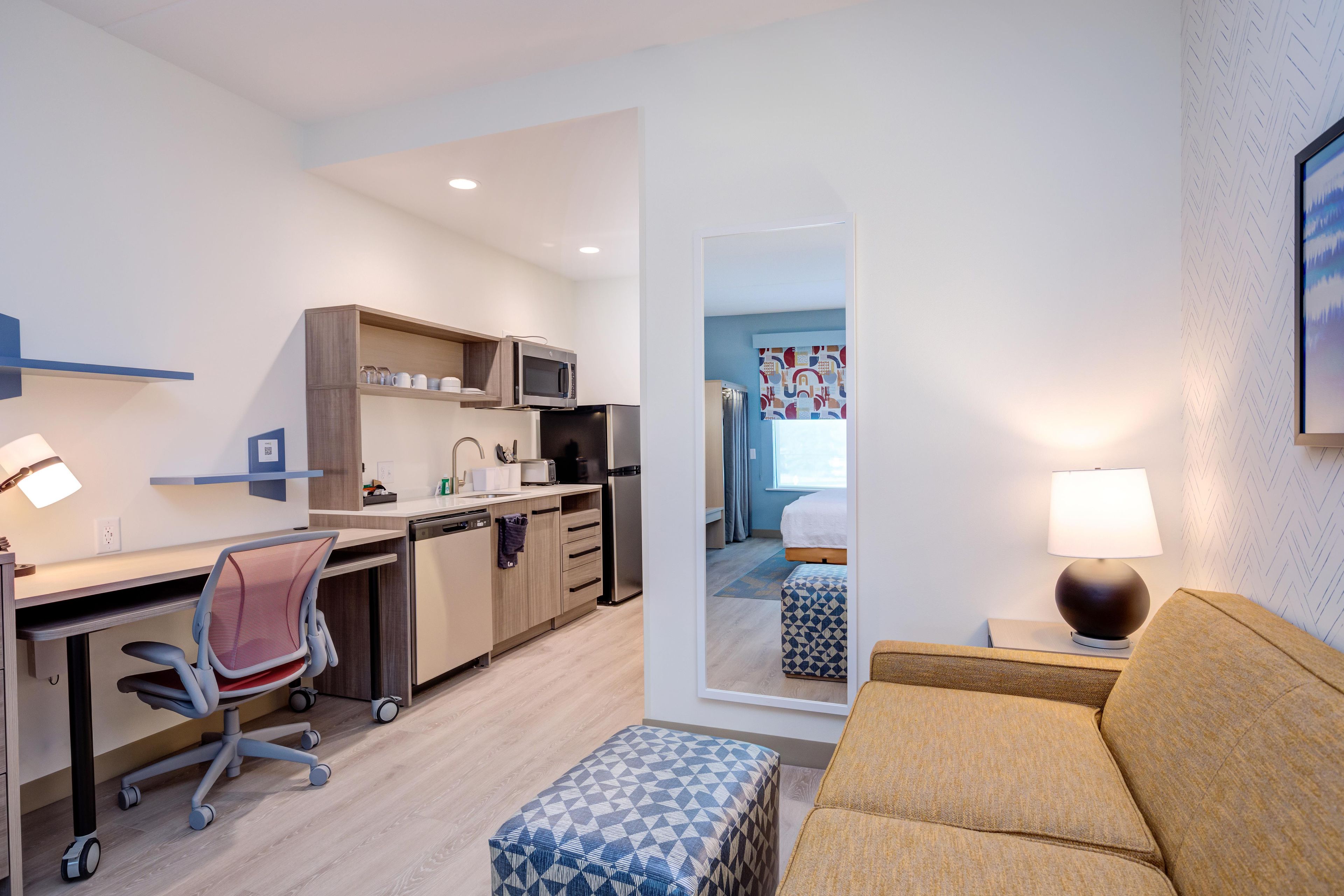 A modern hotel room with a kitchenette, a desk with a chair, a sofa, and a small ottoman.