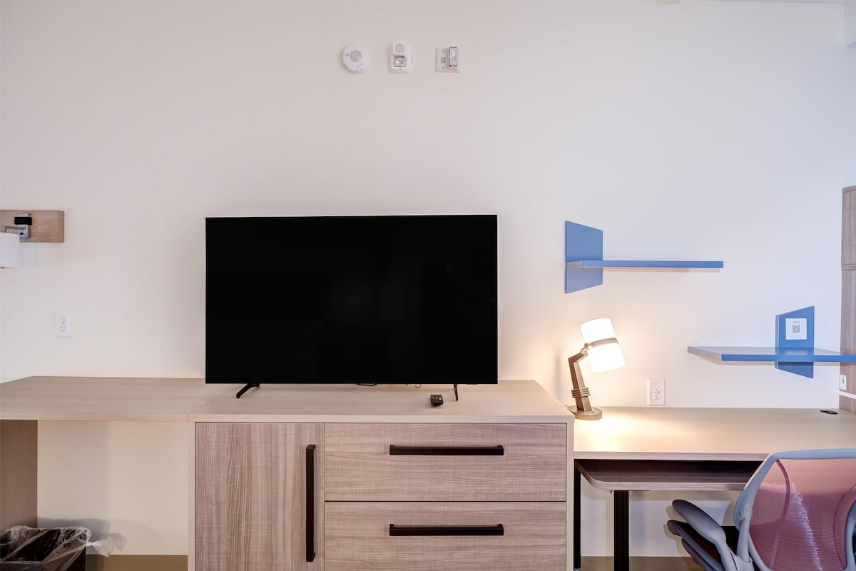 A modern room with a flat-screen TV on a wooden cabinet, a desk with drawers, a lamp, and a blue shelf.