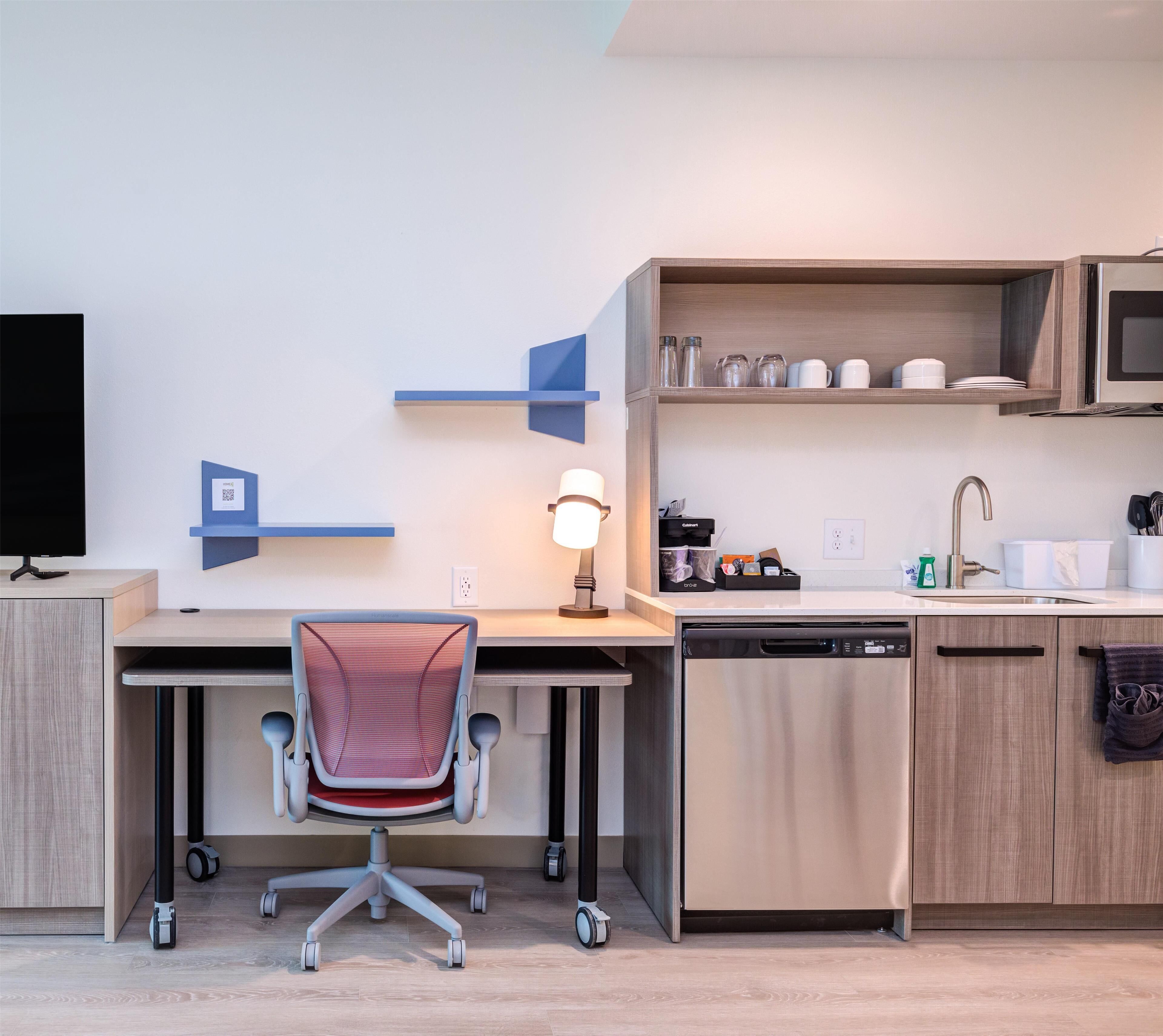 A modern kitchen with a built-in dishwasher, a microwave, and a sink. There is a desk with a chair and a lamp.