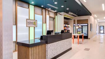 A reception desk with a sign reading "HOME 2 Suites by Hilton," a seating area with a table, and a refrigerator stocked with beverages.