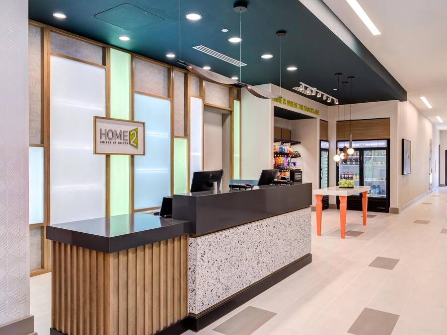A reception desk with a sign reading "HOME 2 Suites by Hilton," a seating area with a table, and a refrigerator stocked with beverages.