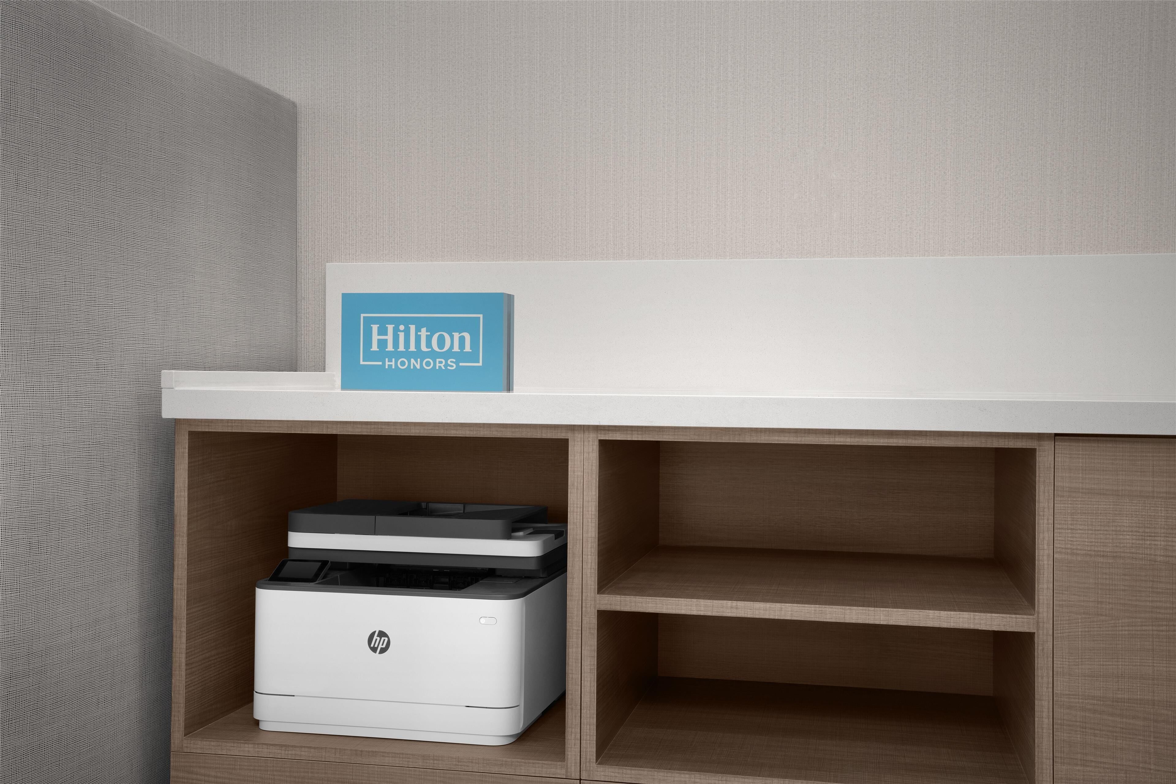 Una camera d'hotel con una scrivania bianca, un cartello Hilton Honors e una stampante HP sulla scrivania.