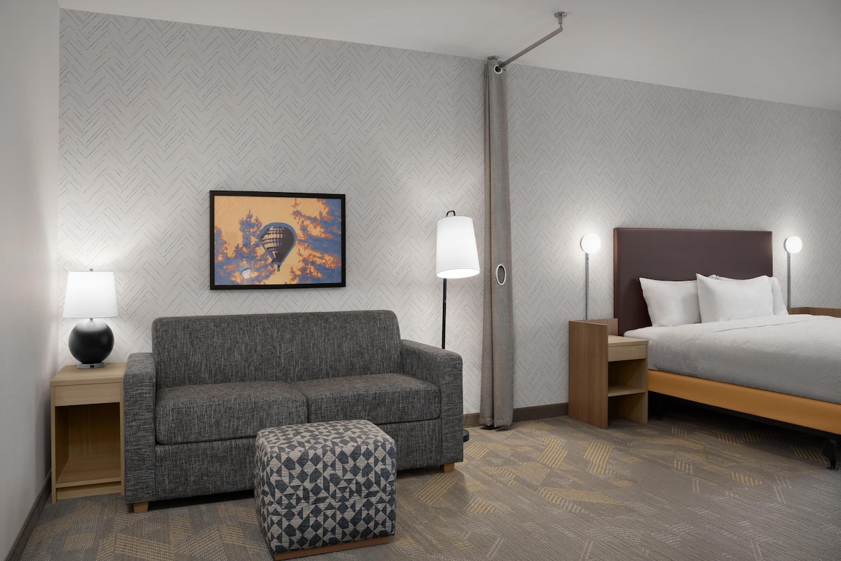A hotel room with a grey sofa, a patterned ottoman, a bed with white bedding, and a wall-mounted artwork.