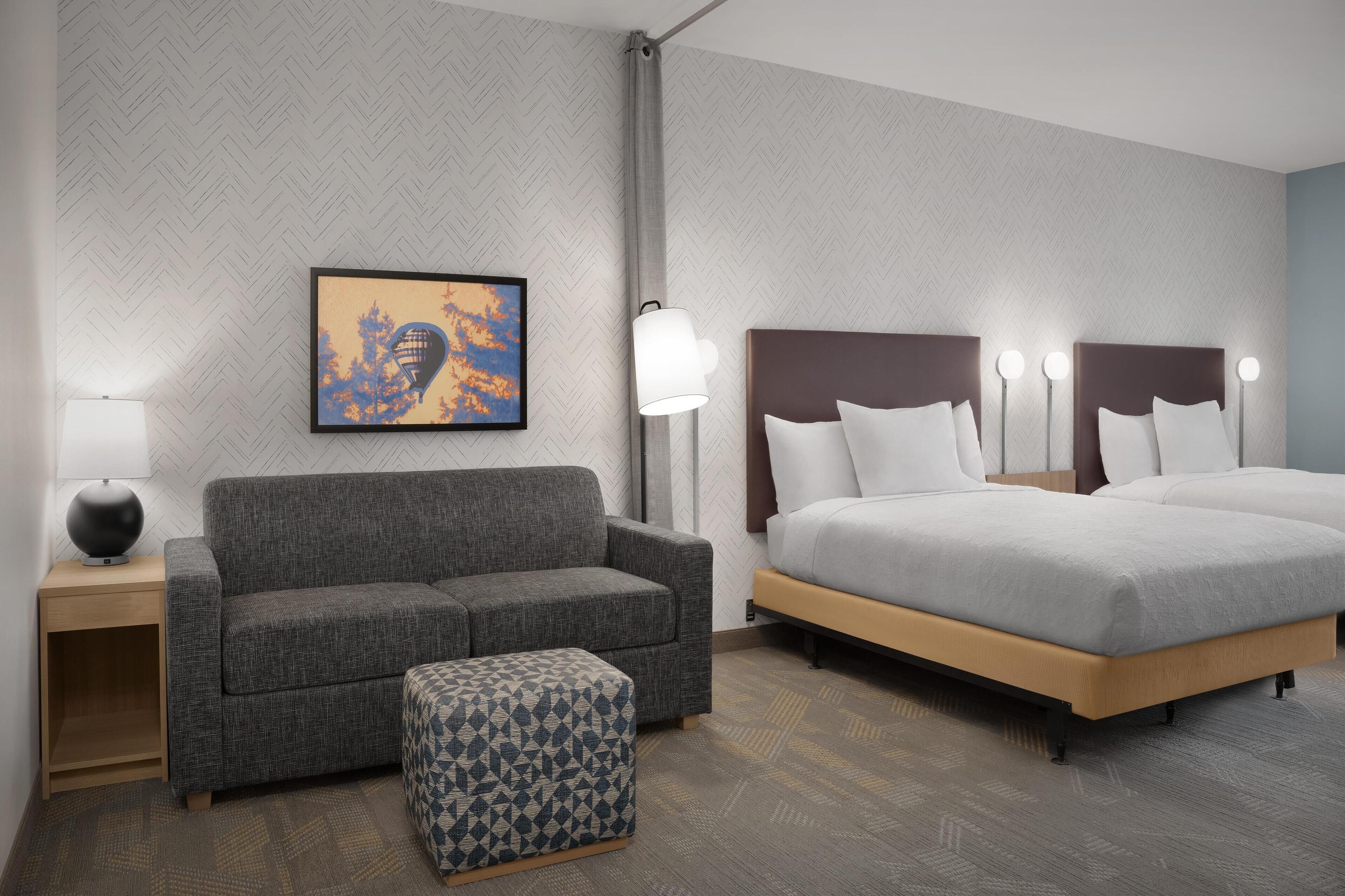 A hotel room with a gray sofa, a patterned ottoman, a large bed with white linens, a nightstand with a lamp, and a framed artwork on the wall.