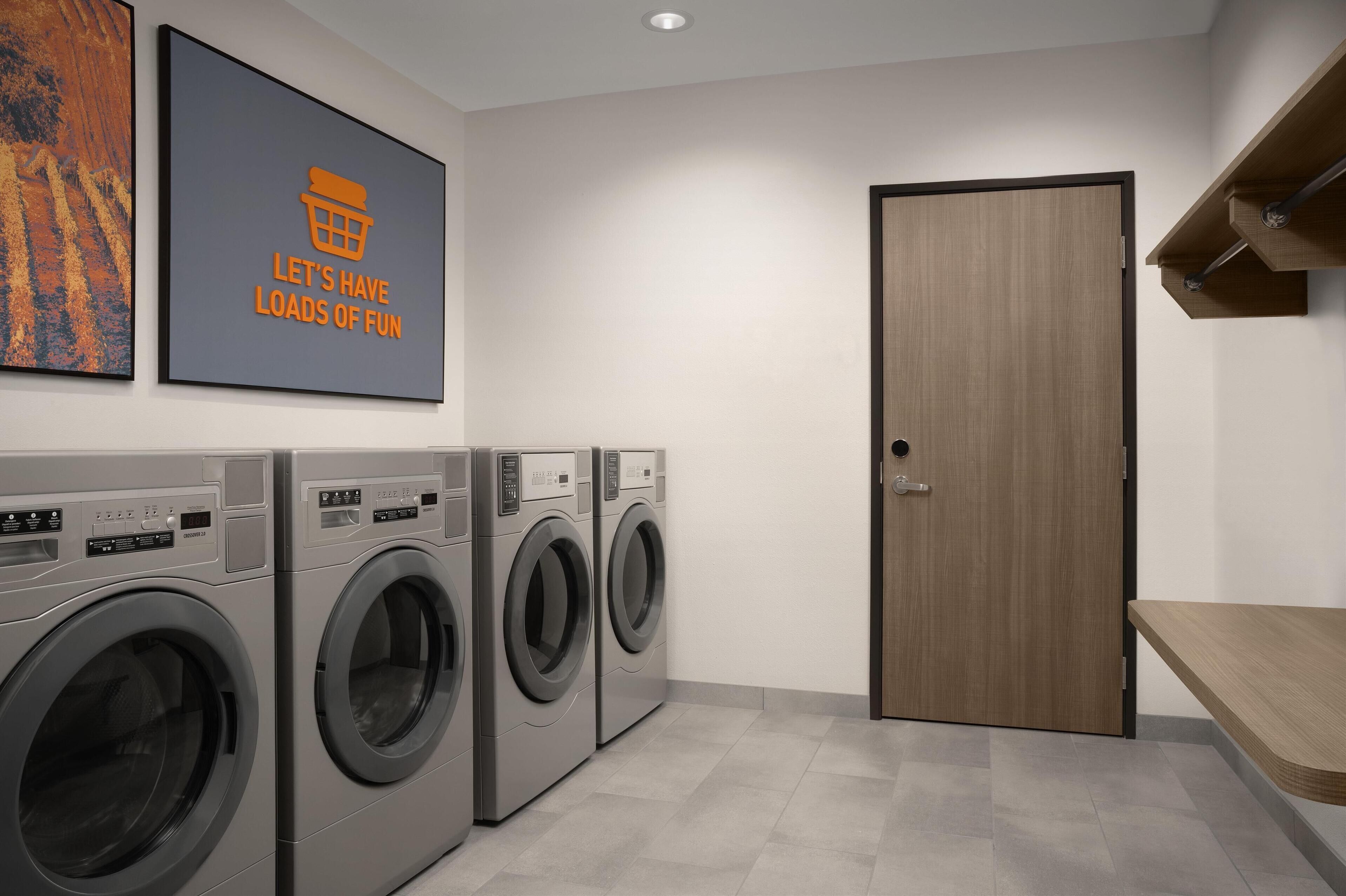 A laundry room with three front-loading washing machines, a wooden door, and a wall-mounted shelf.