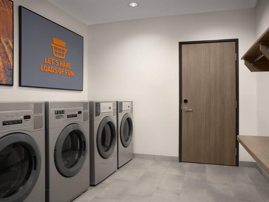A laundry room with three front-loading washing machines, a wooden door, and a wall-mounted shelf.