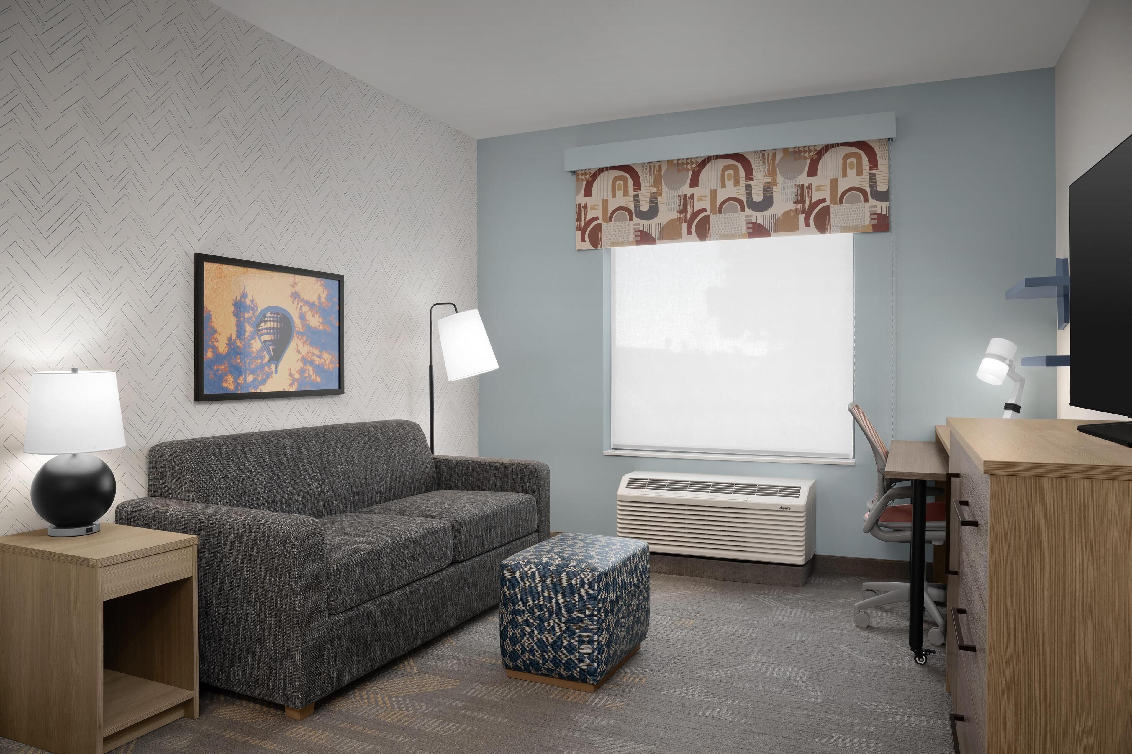 Una sala de estar moderna con un sofá gris, un reposapiés estampado, una mesita de madera con una lámpara, una obra de arte enmarcada, un ventanal con cortina y un escritorio con una silla.