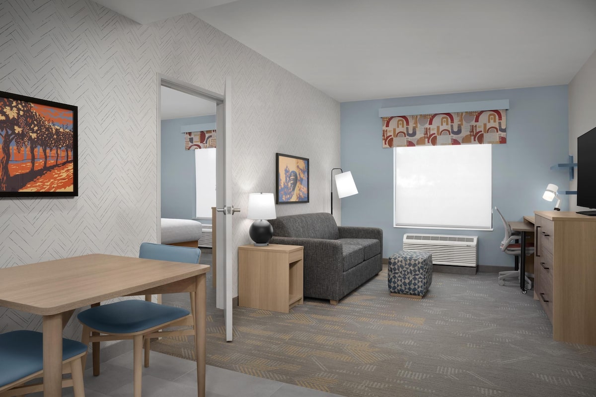 A modern hotel room with a herringbone-patterned wall, a grey sofa, a wooden desk, a television, and a large window with patterned curtains.
