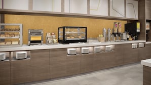 Una barra de desayuno moderna con un sector de autoservicio, que incluye una máquina de café, una vitrina con pasteles y un dispensador de bebidas.