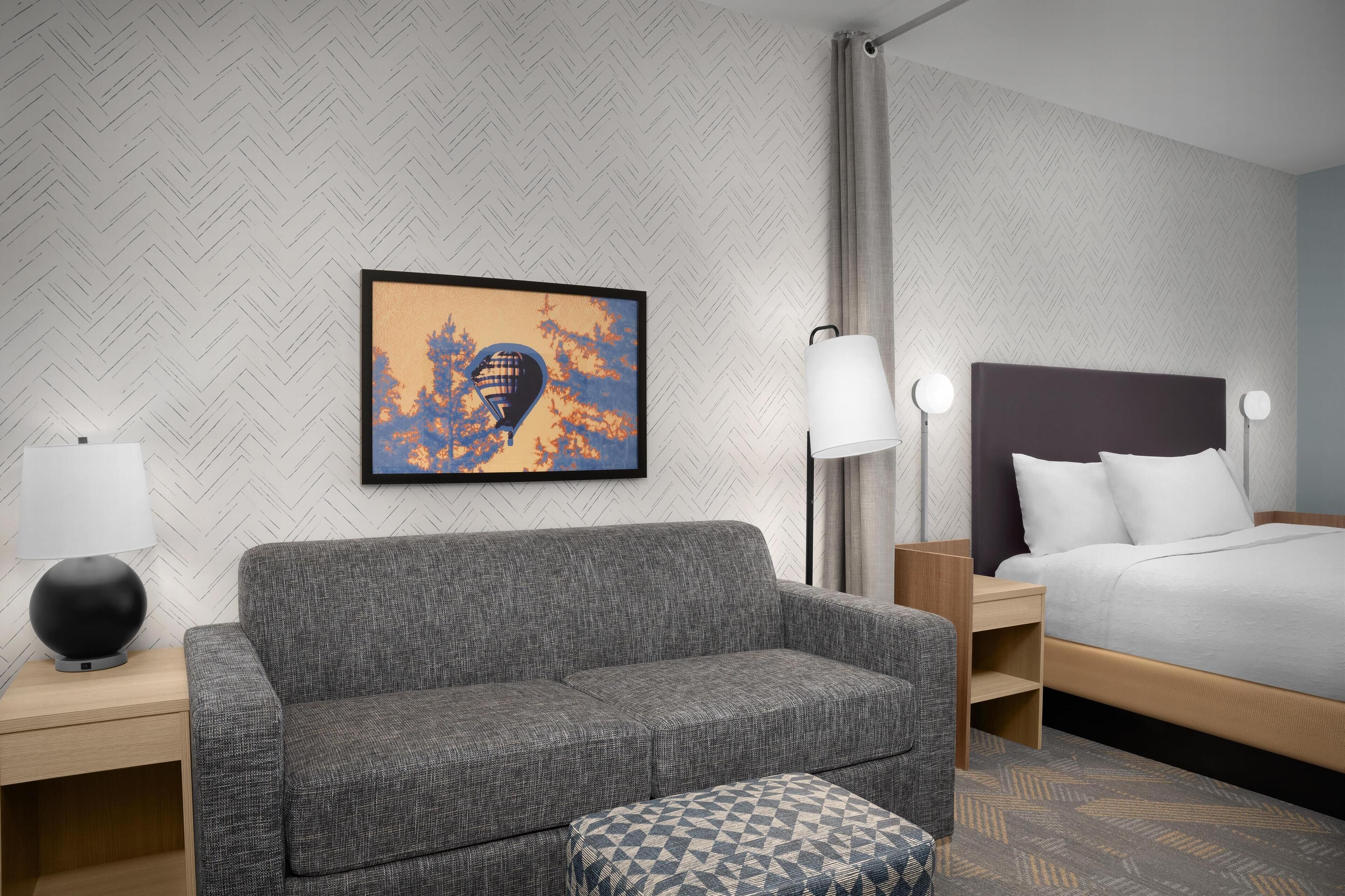 A hotel room with a grey sofa, a patterned ottoman, a bed with white bedding, a nightstand with a lamp, and a framed picture on the wall.