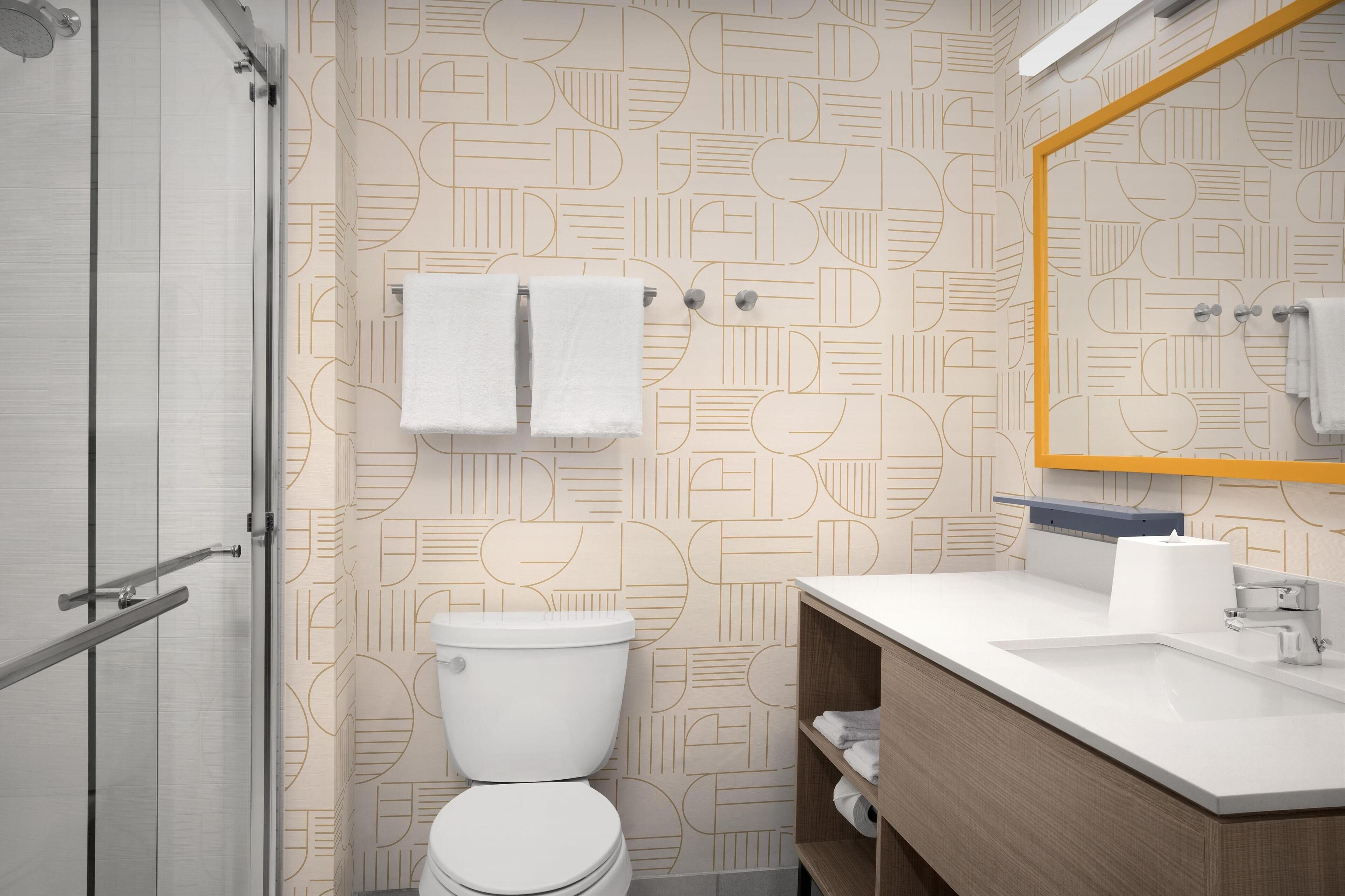 Een moderne badkamer met een wand met een geometrisch patroon, een wit toilet, een houten wastafelmeubel met een spiegel en een glazen douchecabine.