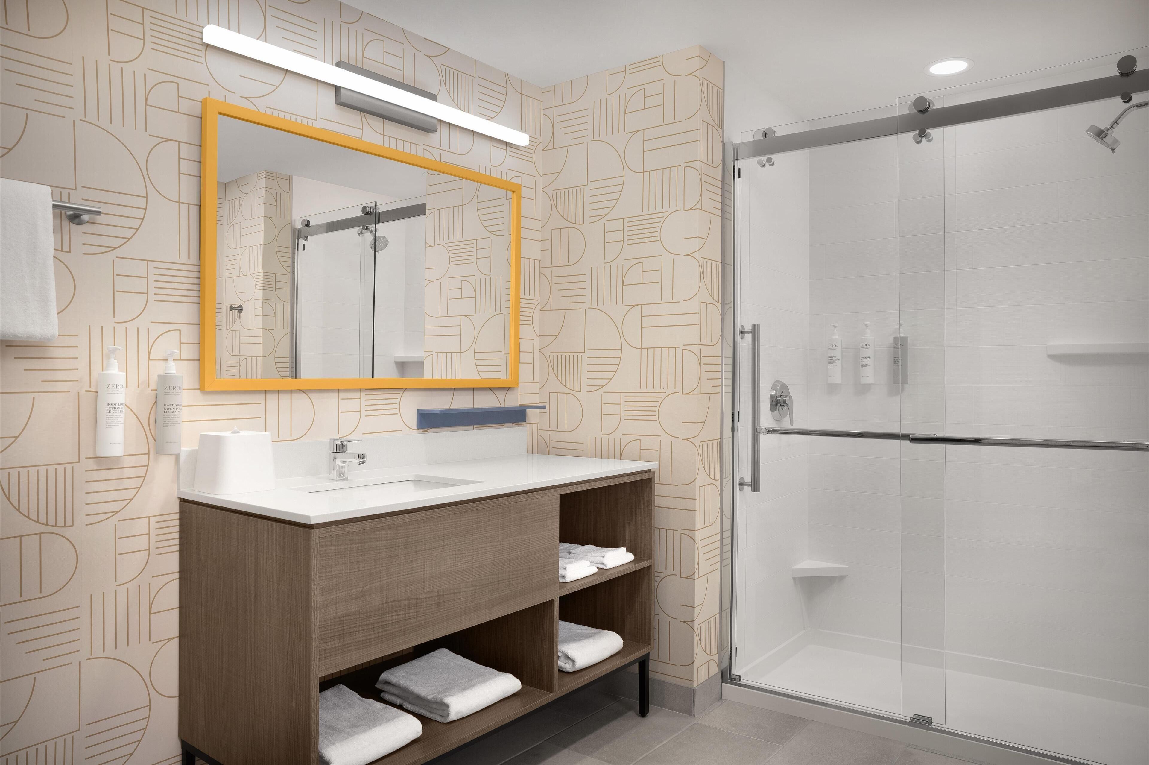 A modern bathroom with a geometric patterned wall, a wooden vanity with a mirror, and a glass shower enclosure.