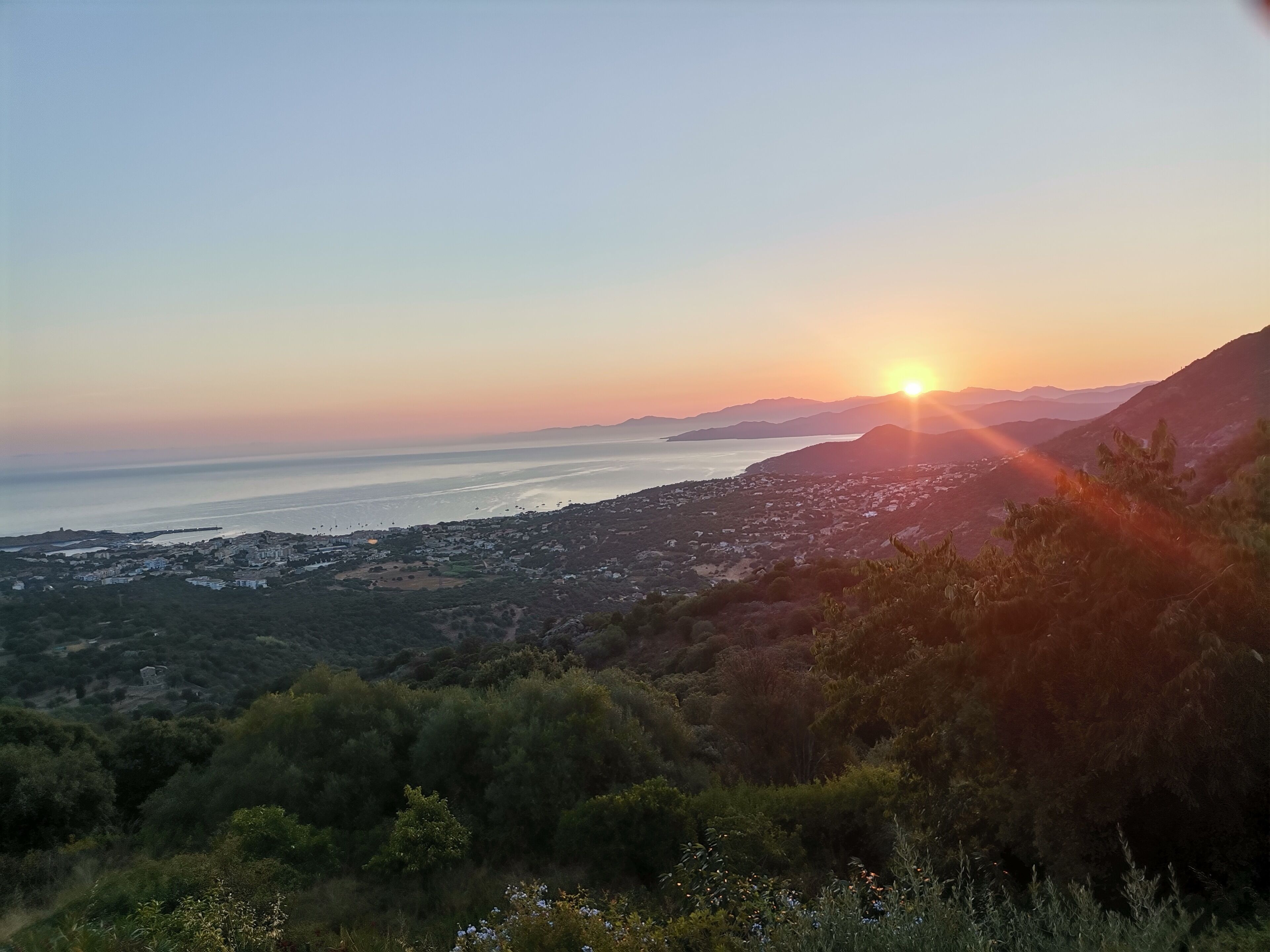 Eine Küstenlandschaft mit Sonnenuntergang, Hügeln und einer Stadt in der Ferne.