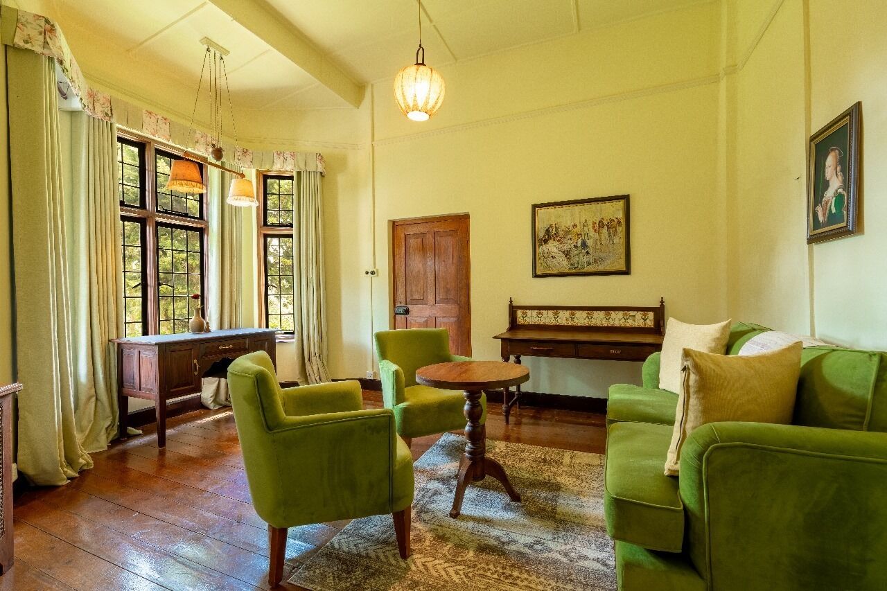 A room with a wooden desk, two green armchairs, a round table, a green sofa, a wooden bench, and a painting on the wall.