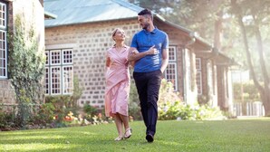 A couple walking on a lawn in front of a stone building with a metal roof. - Bluechip Leisure Summerhill Bungalow (Nuwara Eliya)