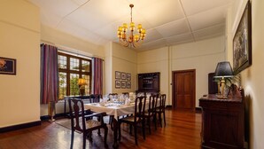 A dining room with a wooden table set for six, surrounded by chairs, a chandelier, and a window with curtains. - Bluechip Leisure Summerhill Bungalow (Nuwara Eliya)